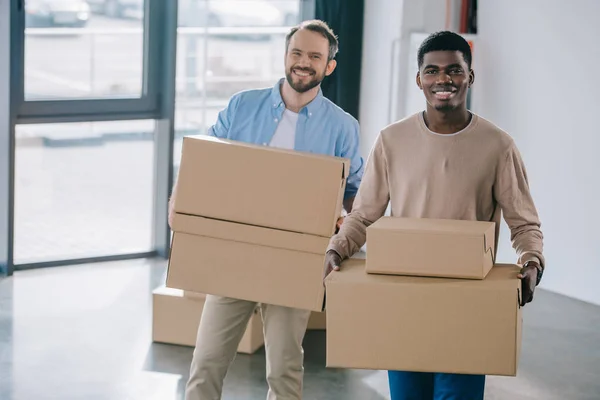 Glückliche multiethnische Männer, die Pappkartons in der Hand halten und beim Umzug in die Kamera lächeln — Stockfoto