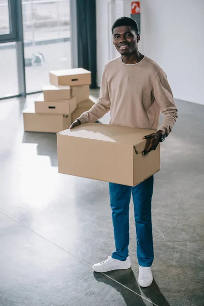 Joven afroamericano hombre sosteniendo caja de cartón y sonriendo a la cámara durante la reubicación - foto de stock