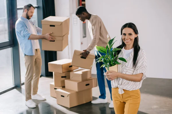 Jeune femme tenant la plante en pot et souriant à la caméra tandis que des collègues masculins tenant des boîtes derrière lors de la relocalisation — Photo de stock