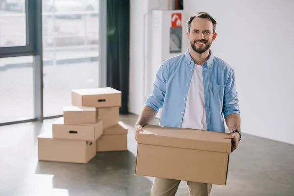 Happy bearded businessman holding cardboard box and smiling at camera while moving in new office — Stock Photo