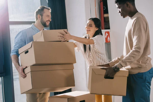 Compañeros de trabajo multiétnicos sonrientes que llevan cajas de cartón durante la reubicación en una nueva oficina - foto de stock
