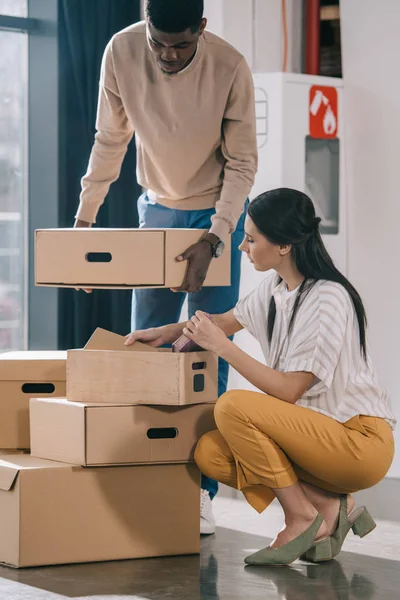 Young multiethnic coworkers unpacking cardboard boxes during relocation in new office — Stock Photo