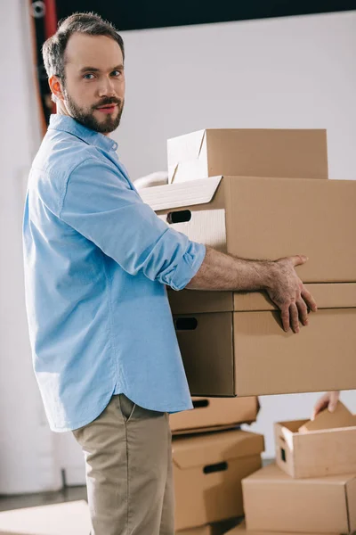 Бородатый бизнесмен держит картонные коробки и смотрит в камеру во время переезда в новый офис — стоковое фото