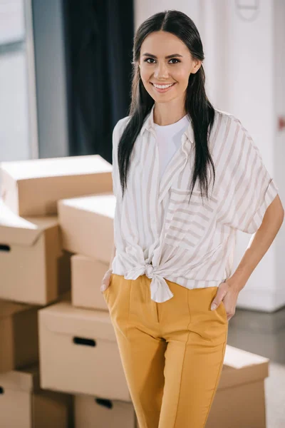 Femme debout avec les mains dans les poches et souriant à la caméra tout en déménageant dans un nouveau bureau — Photo de stock