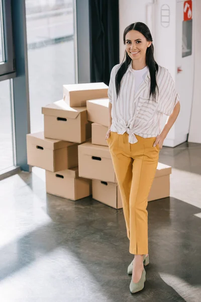 Happy young businesswoman standing with hands in pockets and smiling at camera while relocating in new office — Stock Photo