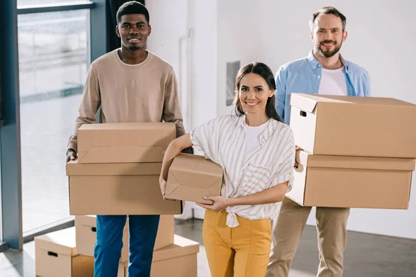 Young multiethnic coworkers holding cardboard boxes and smiling at camera during relocation — Stock Photo
