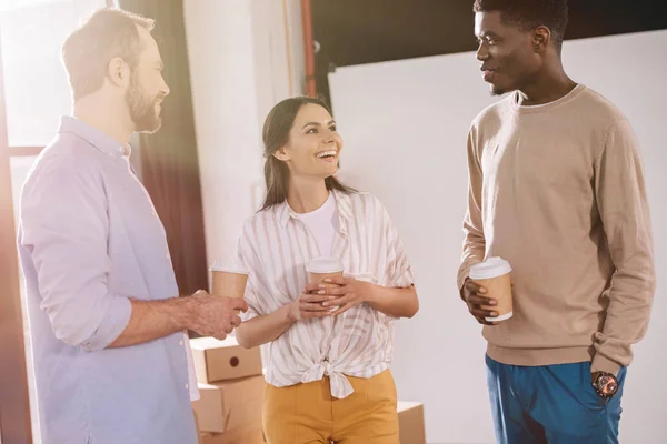 Happy multiethnic coworkers holding paper cups and talking in new office during relocation — Stock Photo