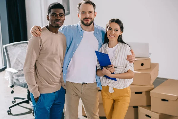 Glückliche multiethnische Kollegen, die im neuen Büro zusammenstehen und in die Kamera lächeln — Stockfoto