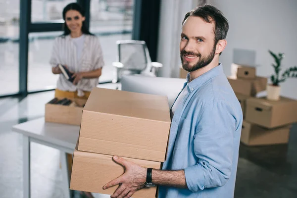 Bel homme barbu tenant des boîtes en carton et souriant à la caméra tout en déménageant avec une collègue féminine dans un nouveau bureau — Photo de stock