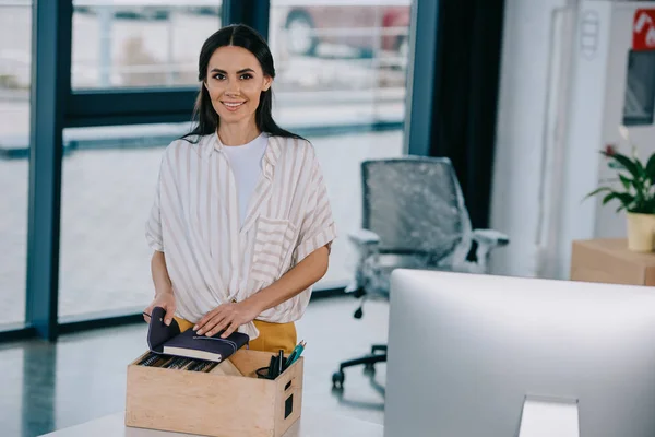 Glückliche junge Geschäftsfrau lächelt in die Kamera, während sie am neuen Arbeitsplatz Büromaterial auspackt — Stockfoto