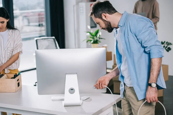 Hombre que sostiene el enchufe de la computadora de escritorio mientras se traslada con sus compañeros de trabajo en nueva oficina - foto de stock