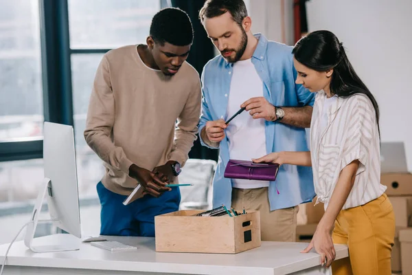 Compañeros de trabajo multiétnicos desempacando caja con suministros de oficina en nuevo lugar de trabajo - foto de stock
