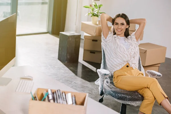 Heureuse jeune femme assise avec les mains derrière la tête et souriant à la caméra dans le nouveau bureau — Photo de stock