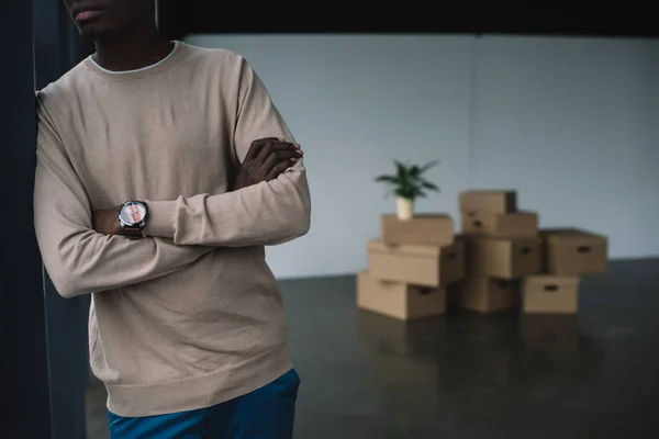 Cropped shot of african american man standing with crossed arms and cardboard boxes behind in new office — Stock Photo