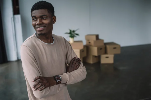 Sorridente afro-americano de pé com braços cruzados e olhando para longe em novo escritório durante a realocação — Fotografia de Stock