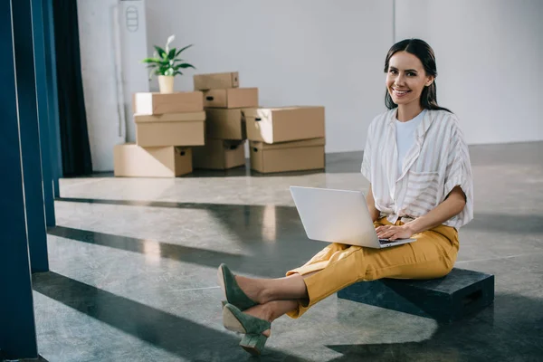 Щаслива молода бізнес-леді використовує ноутбук і посміхається на камеру, сидячи в новому офісі під час переїзду — стокове фото