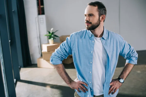 Handsome bearded man standing with hands on waist and looking away during relocation — Stock Photo