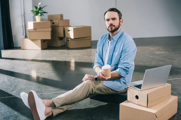 Hombre de negocios sosteniendo café para ir y mirando a la cámara mientras está sentado en el piso en la nueva oficina - foto de stock