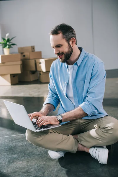 Hombre barbudo sonriente usando el ordenador portátil mientras está sentado en el suelo durante la reubicación - foto de stock