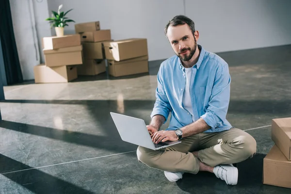 Bel homme barbu à l'aide d'un ordinateur portable et souriant à la caméra tout en étant assis sur le sol dans un nouveau bureau — Photo de stock