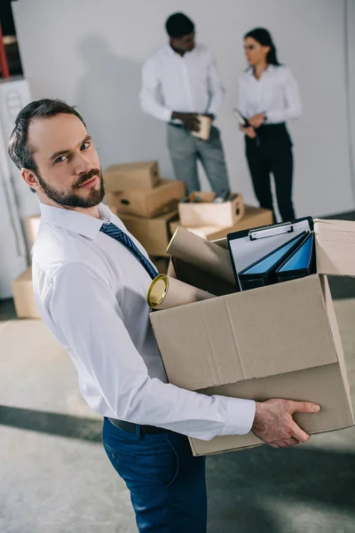 Homme d'affaires tenant une boîte en carton avec des fournitures de bureau et regardant la caméra pendant que des collègues se tiennent derrière dans un nouveau bureau — Photo de stock