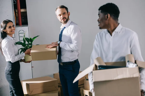 Junger afrikanisch-amerikanischer Geschäftsmann hält Pappschachtel in der Hand und schaut lächelnde Kollegen beim Umzug an — Stockfoto