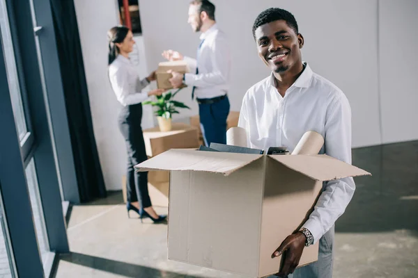 Hombre de negocios afroamericano sosteniendo la caja de cartón y sonriendo a la cámara mientras sus colegas se quedan atrás en una nueva oficina - foto de stock