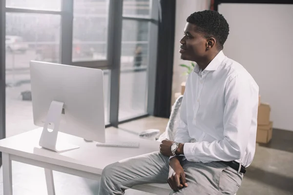 Joven y pensativo empresario afroamericano sentado en la mesa y mirando hacia otro lado en una nueva oficina - foto de stock