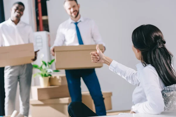 Businesswoman showing thumb up to male colleagues holding cardboard boxes in new office — Stock Photo