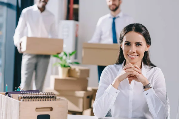 Jovem empresária sentado e sorrindo para a câmera enquanto colegas do sexo masculino segurando caixas atrás no novo escritório — Fotografia de Stock