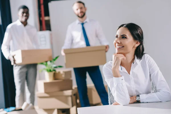 Усміхнена молода бізнес-леді сидить і дивиться далеко, в той час як чоловіки-колеги тримають коробки позаду в новому офісі — стокове фото