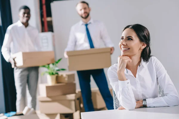 Souriant jeune femme d'affaires assis et regardant loin tandis que des collègues masculins tenant des boîtes derrière dans le nouveau bureau — Photo de stock