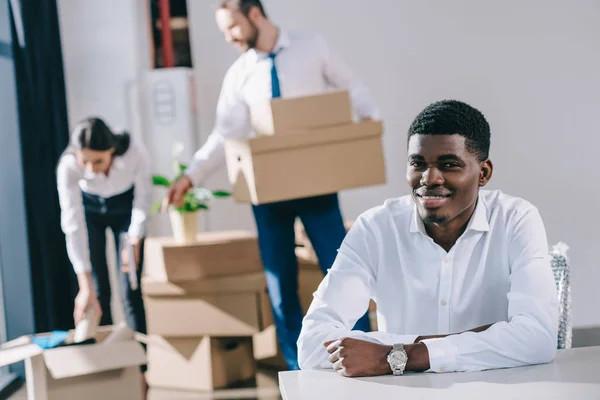 Young african american businessman smiling at camera while colleagues unpacking boxes in new office — Stock Photo