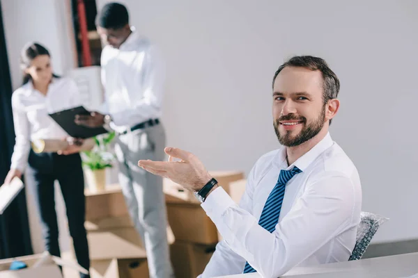 Счастливый бизнесмен улыбается в камеру, в то время как коллеги стоят позади в новом офисе — стоковое фото