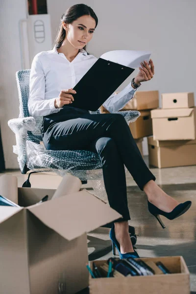 Concentré jeune femme d'affaires tenant presse-papiers tout en étant assis dans un nouveau bureau pendant la réinstallation — Photo de stock