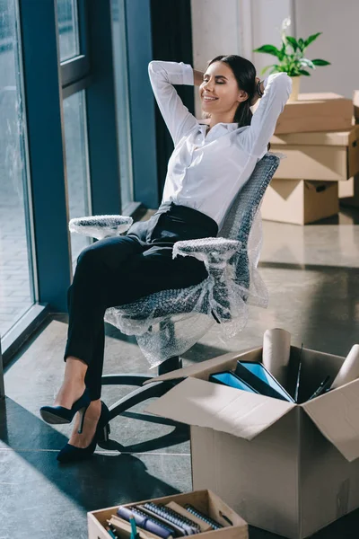 Счастливая молодая деловая женщина сидит с руками за головой в новом офисе — стоковое фото
