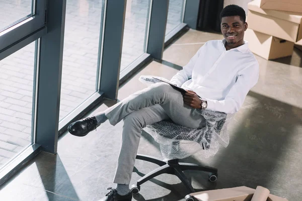 Jeune homme d'affaires afro-américain avec presse-papiers assis et souriant à la caméra dans un nouveau bureau — Photo de stock
