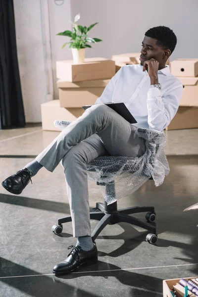 Pensive young african american businessman with clipboard sitting and looking away in new office — Stock Photo