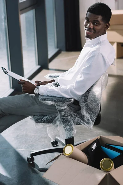 Vista de ángulo alto del hombre de negocios afroamericano sujetando el portapapeles y sonriendo a la cámara mientras está sentado en una nueva oficina durante la reubicación - foto de stock