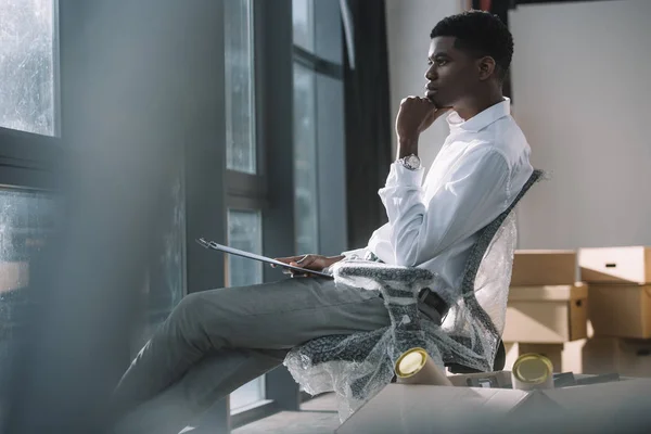 Молодий афроамериканський бізнесмен тримає кишеню і дивиться на вікно, сидячи в новому офісі під час переїзду — стокове фото
