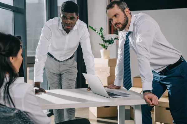 Hombres de negocios multiétnicos apoyados en la mesa y mirando a la mujer de negocios en una nueva oficina - foto de stock