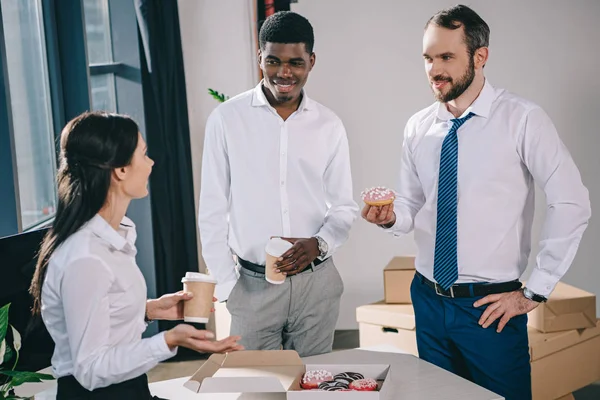 Sonrientes compañeros de trabajo multiétnicos bebiendo café para ir a comer rosquillas en una nueva oficina - foto de stock