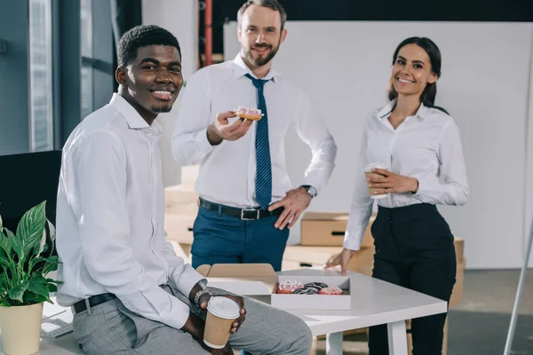 Compañeros de trabajo multiétnicos felices sosteniendo tazas de papel con café para ir y sonriendo a la cámara en la nueva oficina - foto de stock