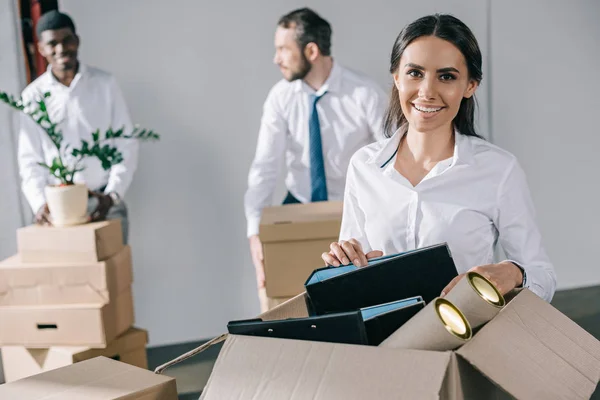 Glückliche junge Geschäftsfrau packt Karton mit Büromaterial aus und lächelt in die Kamera, während männliche Kollegen im neuen Büro hinten stehen — Stockfoto