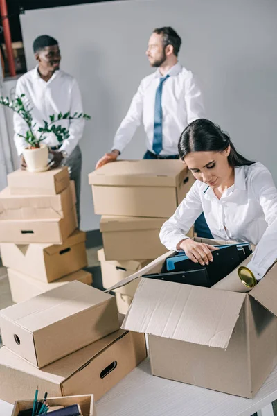 Young businesswoman unpacking cardboard box and multiethnic men standing behind in new office — Stock Photo
