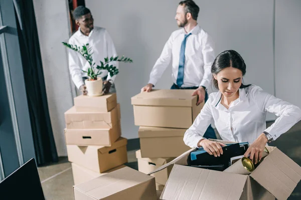 Jeune femme d'affaires déballage boîte en carton et collègues masculins debout derrière dans le nouveau bureau — Photo de stock