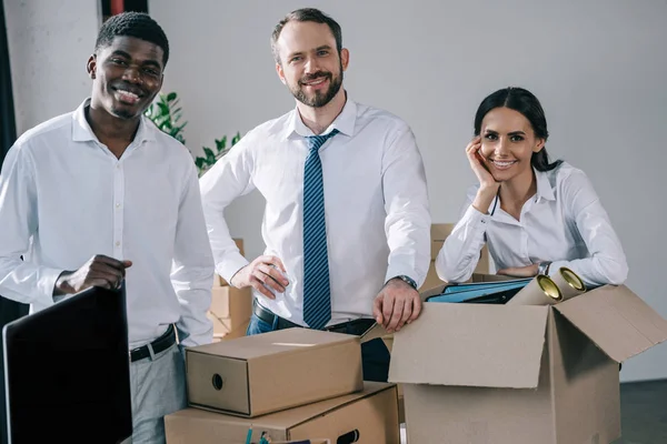 Glückliche multiethnische Kollegen lächeln in die Kamera, während sie im neuen Büro Kartons auspacken — Stockfoto