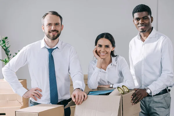 Heureux collègues multiraciaux souriant à la caméra et déballer les boîtes dans le nouveau bureau — Photo de stock