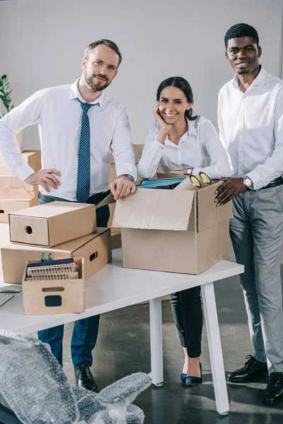 Happy multiethnic coworkers smiling at camera and unpacking boxes in new office — Stock Photo