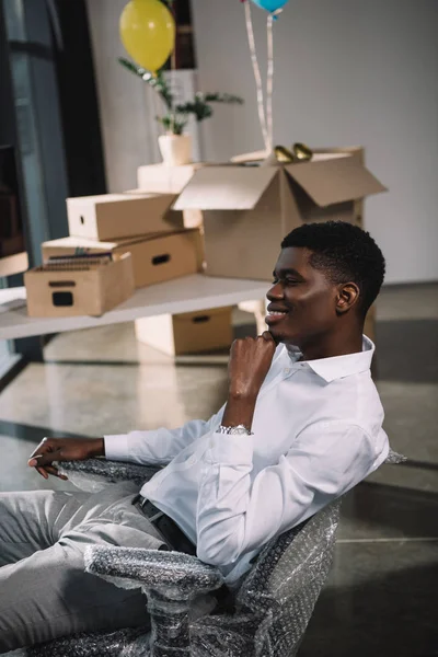 Sonriente joven hombre de negocios afroamericano sentado en el lugar de trabajo en una nueva oficina - foto de stock